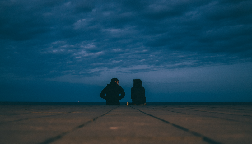 A couple sitting apart on a dimly lit promenade under a cloudy sky, symbolizing relationship difficulties and uncertainty, which may impact a spouse visa situation.