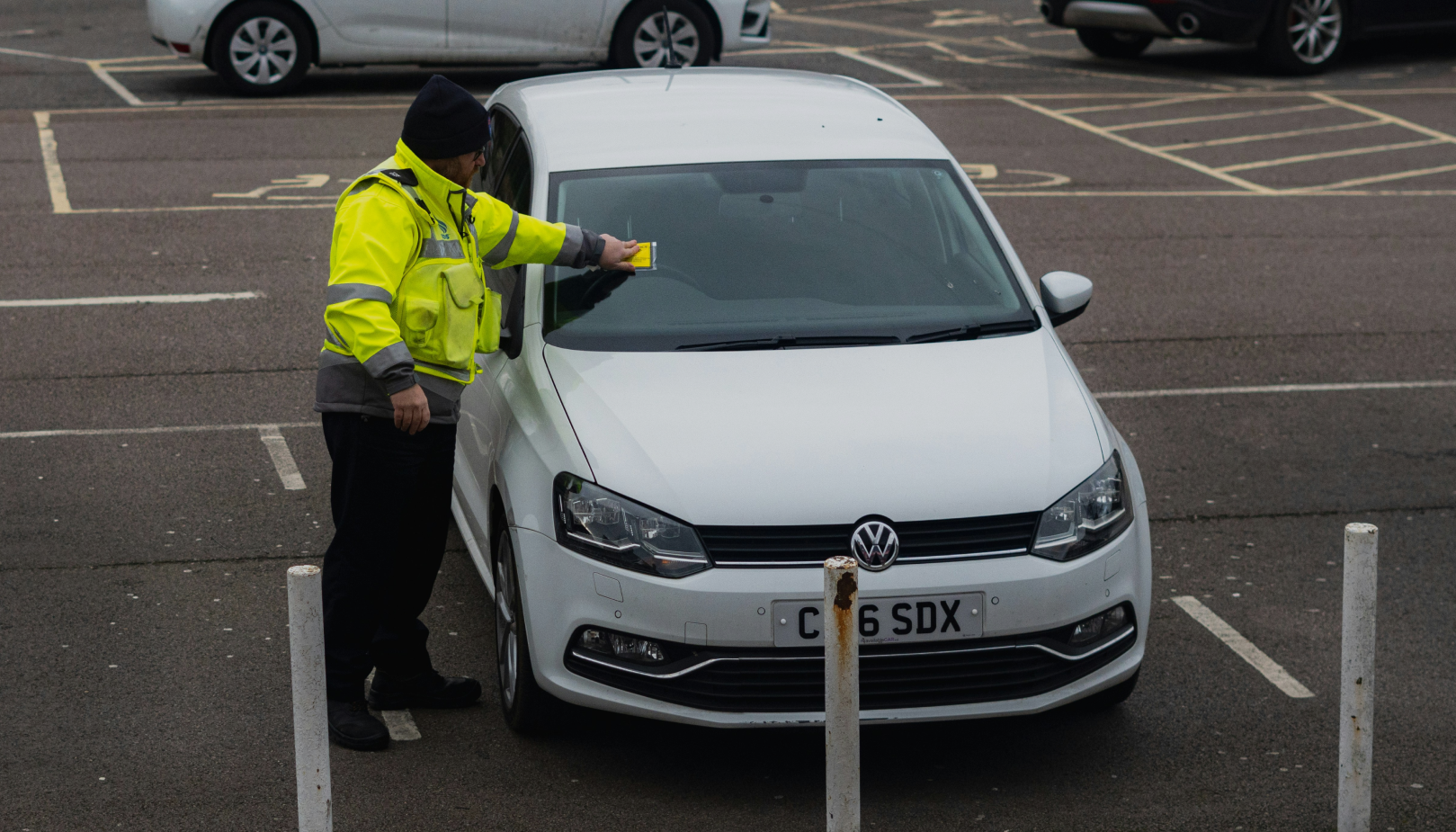 Parking enforcement officer in a high-visibility jacket placing a penalty notice on the windshield of a white Volkswagen in a parking lot.
