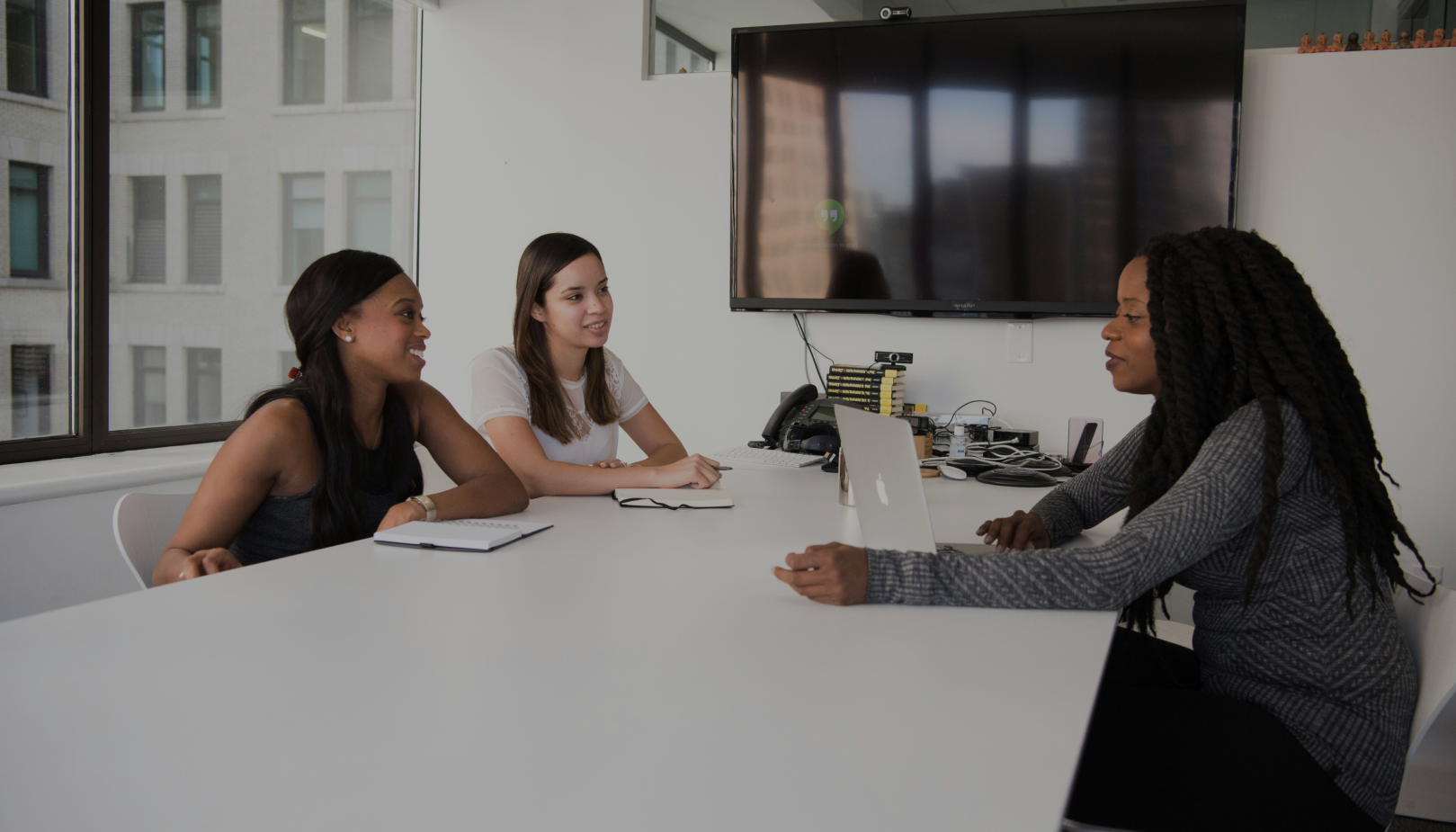 Three women having a discussion in a modern office conference room with a laptop and notepad on the table.