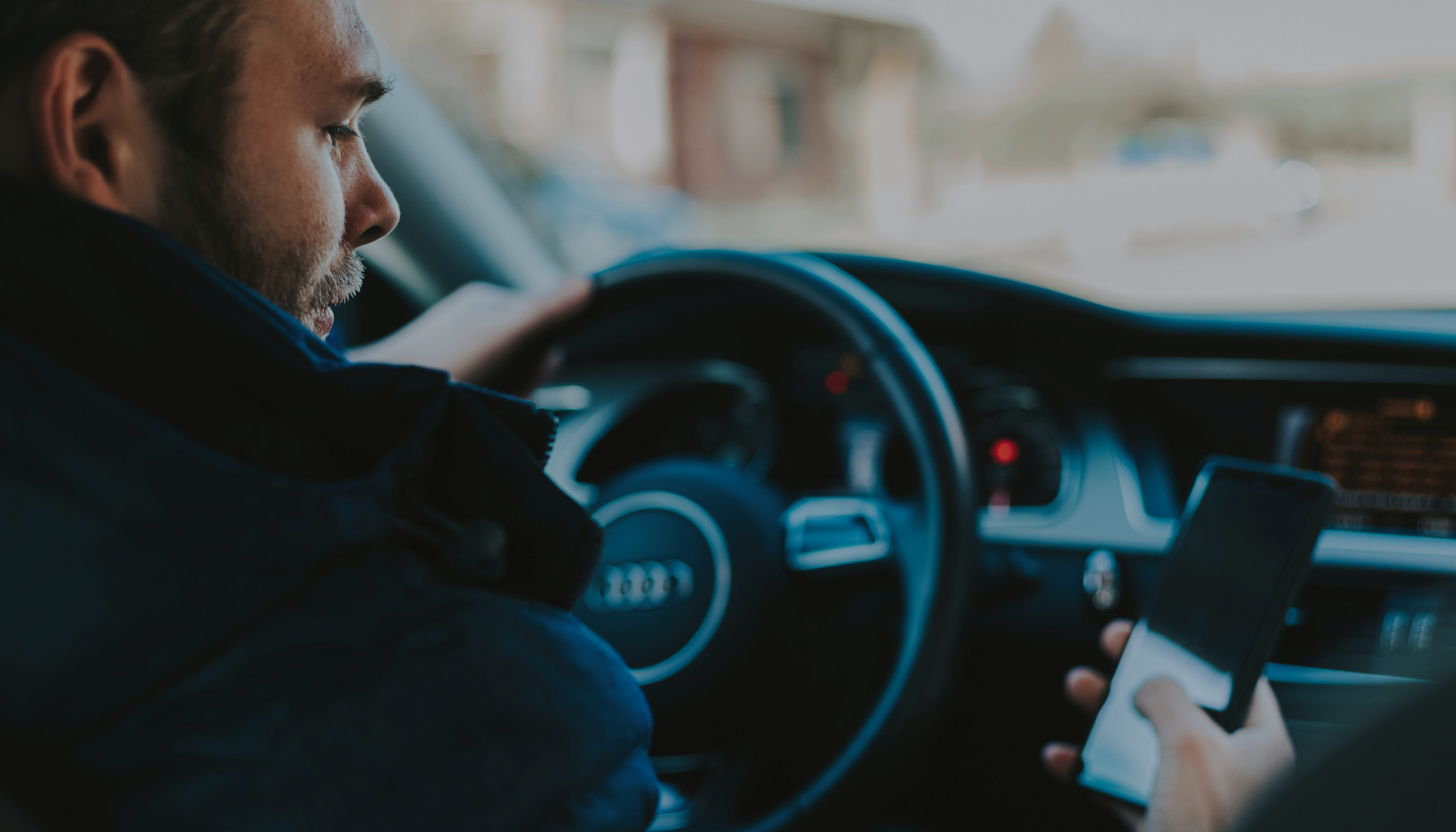 Man driving an Audi while looking at his smartphone, with one hand on the steering wheel and the other holding the phone.