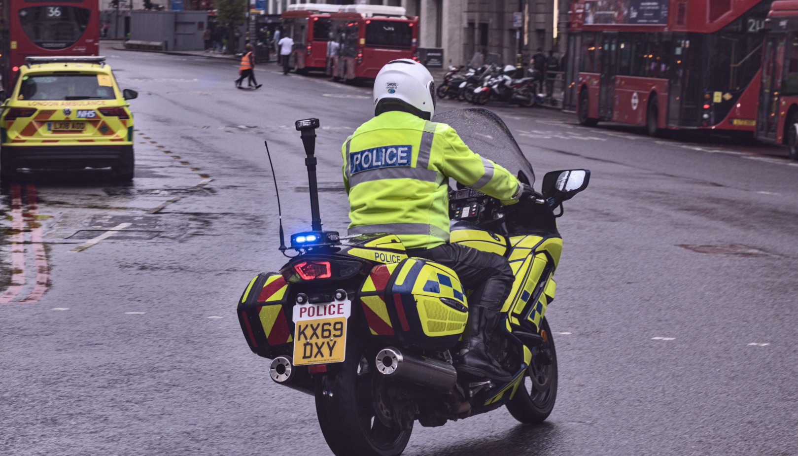 UK police officer on a high-visibility motorcycle riding through a wet city street, with emergency lights on and red double-decker buses in the background.