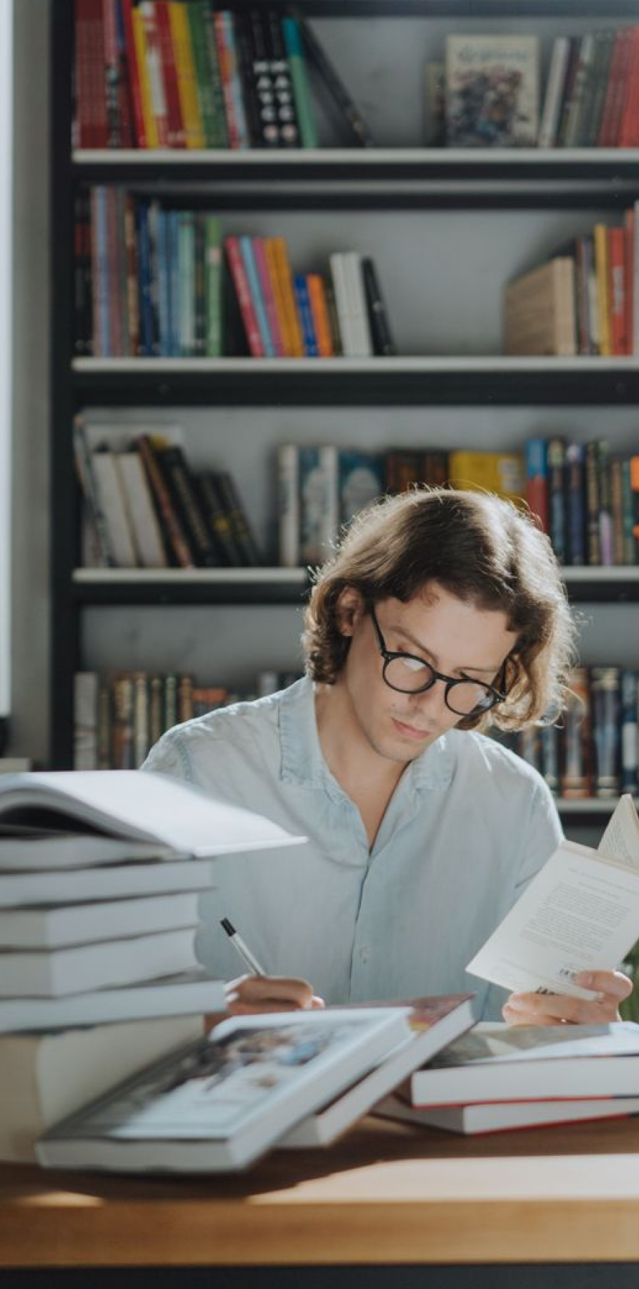 A man wearing glasses is engrossed in a book, seated in front of a filled bookcase